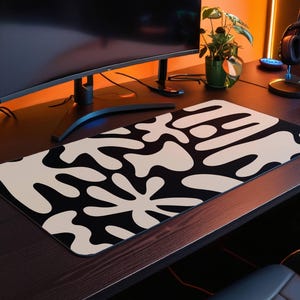 May include: A black and white abstract design mousepad with large white shapes on a black background. The mousepad is on a wooden desk with a computer monitor, a plant, and a lamp.