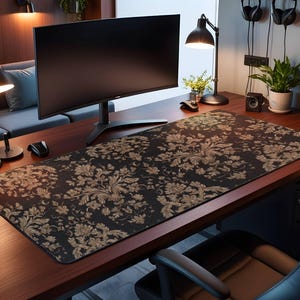 Puede incluir: Una alfombrilla de escritorio con estampado floral negro y dorado sobre un escritorio de madera oscura. Un monitor de ordenador negro, una lámpara de escritorio negra y plantas en macetas también están en el escritorio. Una silla de oficina negra está frente al escritorio.