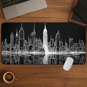 Könnte beinhalten: Schwarze rechteckige Schreibtischunterlage mit einer weißen Strichzeichnung einer Skyline und Reflexionen. Das Empire State Building ist prominent. Eine weiße Computermaus liegt auf der Unterlage. Die Unterlage liegt auf einem Holzschreibtisch.