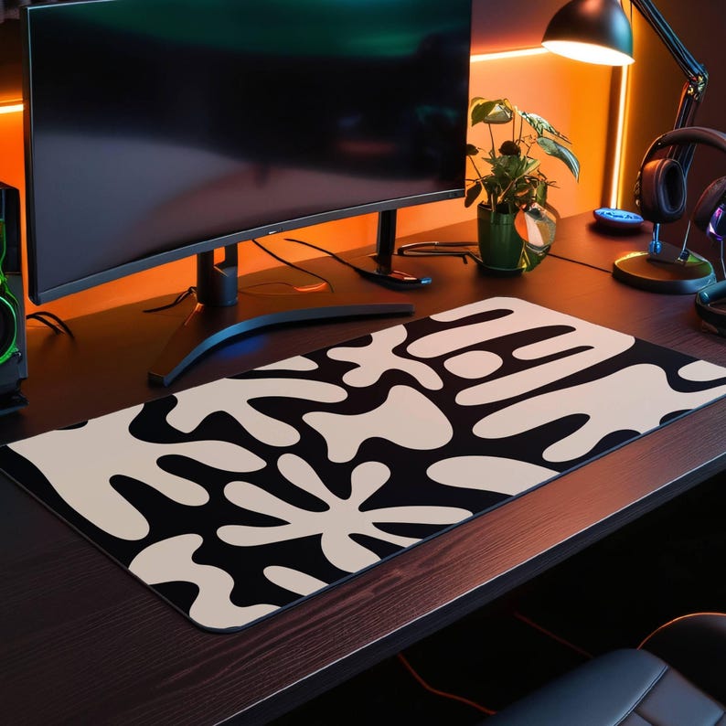 May include: A black and white abstract design mousepad with large white shapes on a black background. The mousepad is on a wooden desk with a computer monitor, a plant, and a lamp.