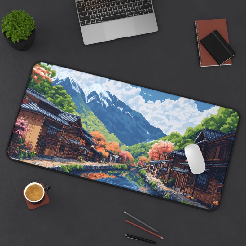 Pixel art desk mat of a traditional Japanese village with wooden buildings, a canal, cherry blossom trees, and snow-capped mountains in the background. The scene is colorful, with reflections in the water, a blue sky, and green hills.