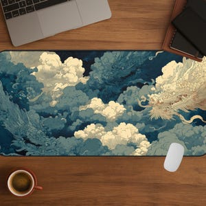 Puede incluir: Alfombrilla de escritorio rectangular con una ilustración detallada de dos dragones entre nubes arremolinadas. Los dragones están representados en tonos azules y crema, sobre un fondo de nubes azules y blancas. Se ve un ratón de ordenador y una taza de café.