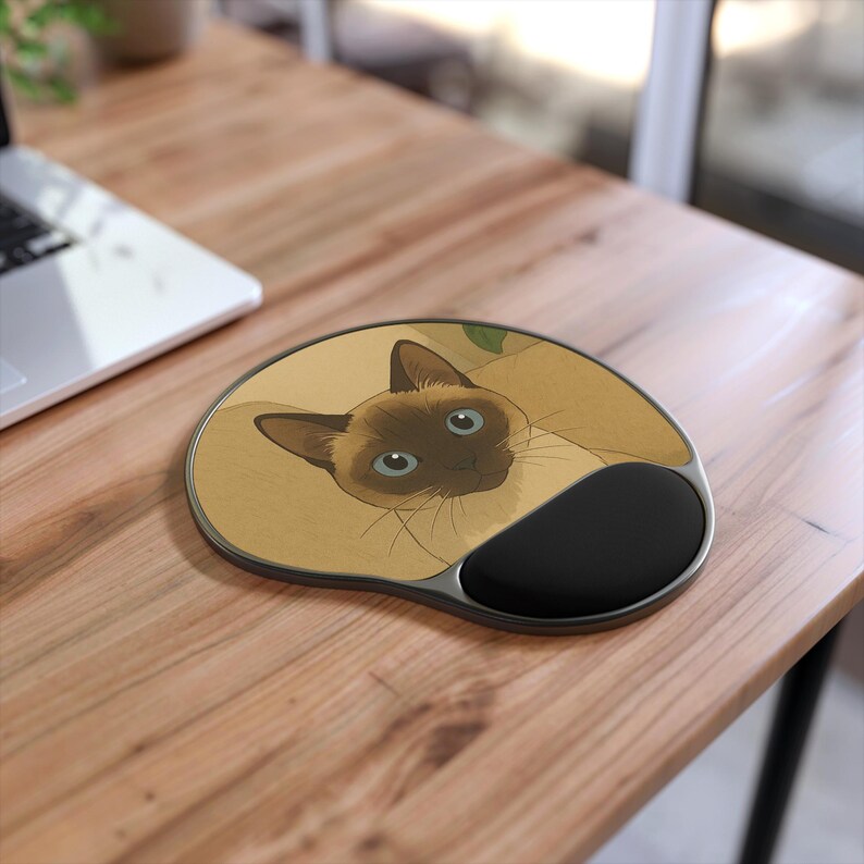 Pode incluir: Um mousepad ergon&oacute;mico bege e preto com uma ilustra&ccedil;&atilde;o de um gato siam&ecirc;s. O gato tem pelo castanho, olhos azuis e um nariz preto. O mousepad tem um apoio de pulso preto e uma borda prateada. Numa secret&aacute;ria de madeira.
