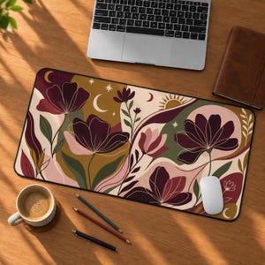 May include: A rectangular desk mat featuring a floral pattern with dark purple, pink, olive green, and mustard yellow shapes. The design includes celestial elements like crescent moons, a sun, and stars against a cream-colored background.