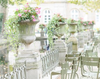 May include: A row of white stone urns with pink flowers and green foliage, set against a white building with windows.  The urns are lined up along a path with a wrought iron fence and green metal chairs.