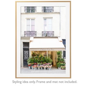 May include: A framed print of a Parisian flower shop, featuring a cream-colored awning with the text "L'histoire d'une fleur." The shop's facade is a light beige, with windows and wrought-iron balconies above. Colorful flowers are displayed in buckets.