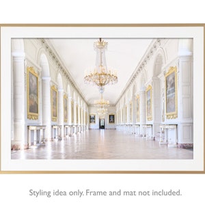 May include: A photograph of a long, white hallway with a large chandelier hanging from the ceiling. The hallway is lined with arched doorways and paintings on the walls. The floor is tiled in a light colour.