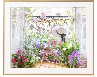 Op de afbeelding: Een roze metalen stoel met een witte hoed op de rugleuning, omringd door verschillende bloemen in roze, wit en paarse tinten. De stoel staat voor een raam met uitzicht op een balkon en een smeedijzeren balustrade.