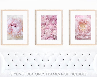 May include: Three framed floral prints featuring close-up images of pink peonies. The prints are displayed above a white tufted sofa. The frames are a light wood color, and the flowers have varying shades of pink and white.
