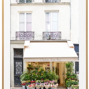 May include: A framed photograph of a Parisian flower shop. The shop has a cream-colored awning with the text "L'histoire d'une fleur." The shop's facade is cream-colored, with black wrought-iron balconies and a black door. A variety of flowers are displayed in buckets.