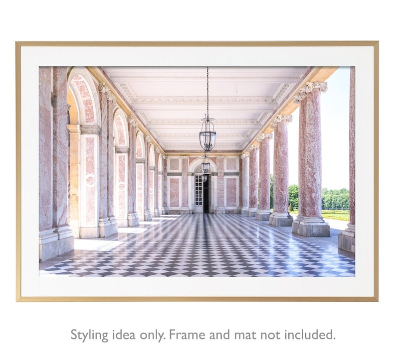 May include: A framed print of a classical colonnade with pink marble columns and arched openings. The black and white checkered floor leads to a distant doorway. Two ornate hanging lanterns illuminate the space. The frame is gold.