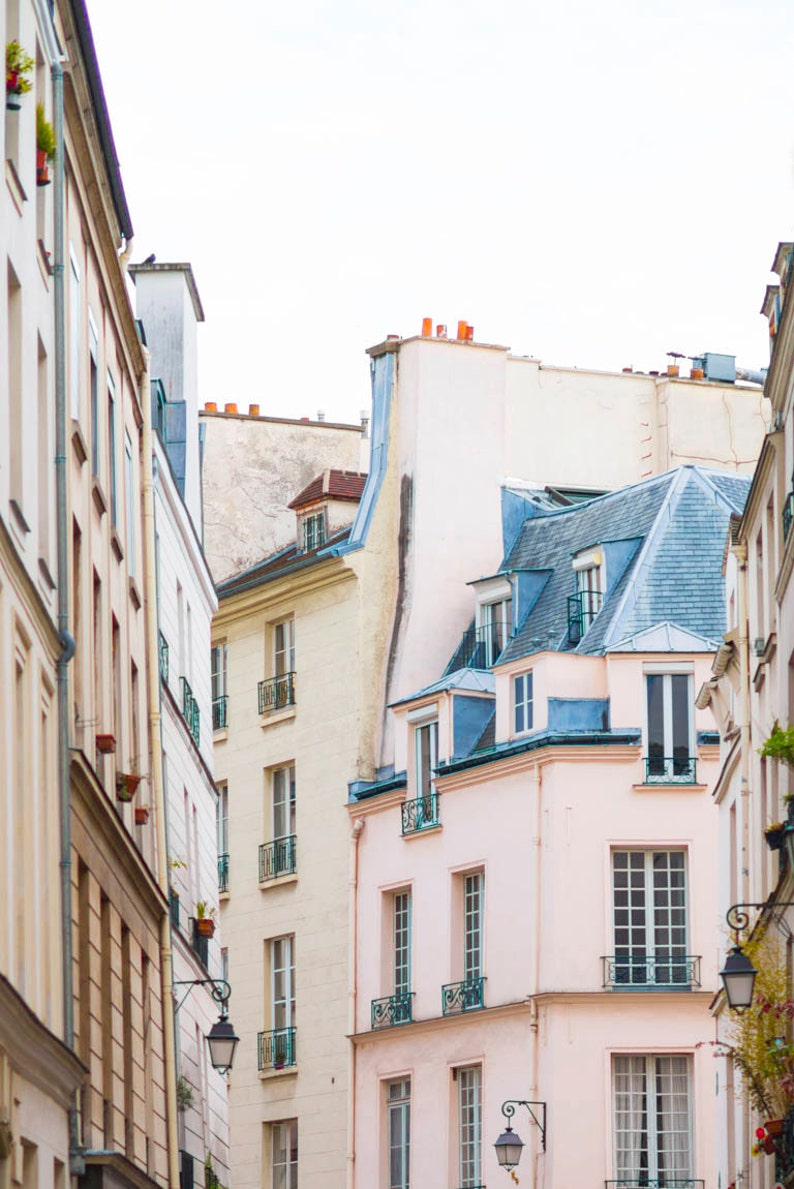May include: A narrow street in Paris, France, with a row of old buildings. The buildings are painted in shades of white, beige, and pink. The buildings have traditional French architecture, with dormer windows and decorative details. The street is lined with lampposts.