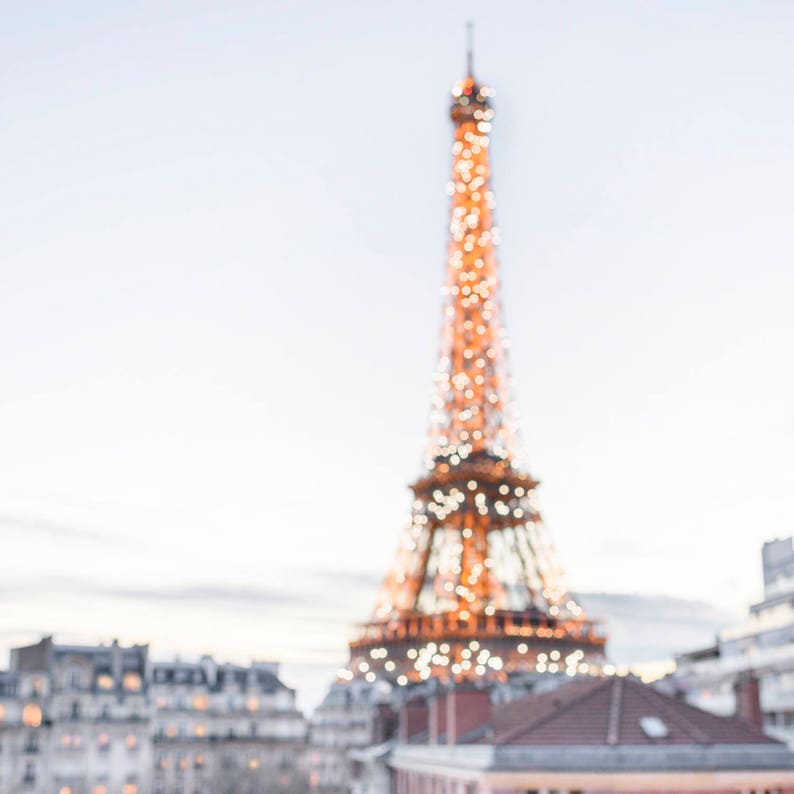May include: A blurry image of the Eiffel Tower in Paris, France, with lights on at dusk. The tower is in the background, and the foreground shows the rooftops of buildings in the city.