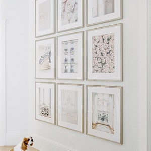 May include: A gallery wall with nine framed prints in a white room. The prints feature various images, including flowers, architecture, and a staircase. The frames are all the same size and have a light brown finish. The wall is painted white and the floor is a light wood.