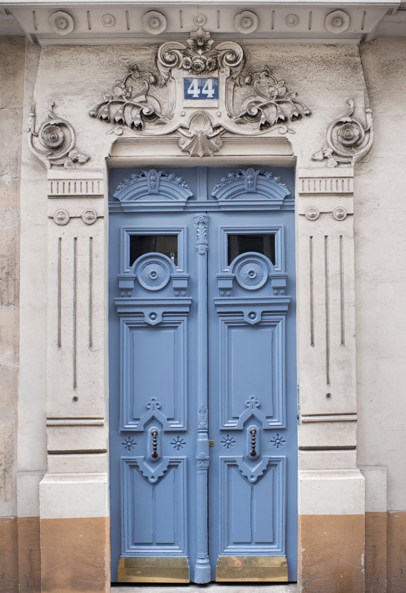 May include: A blue double door with ornate details and a brass door handle. The door is framed by a white stone archway with a decorative stone plaque above the door that reads "44".