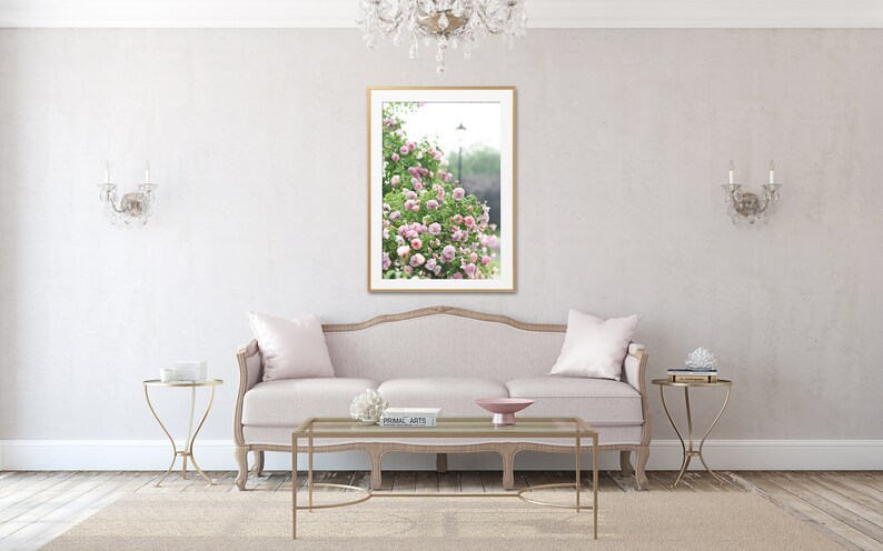 May include: A living room with a light pink sofa, gold side tables, and a framed picture of pink flowers. The room has a neutral colour palette with a rug and a chandelier.