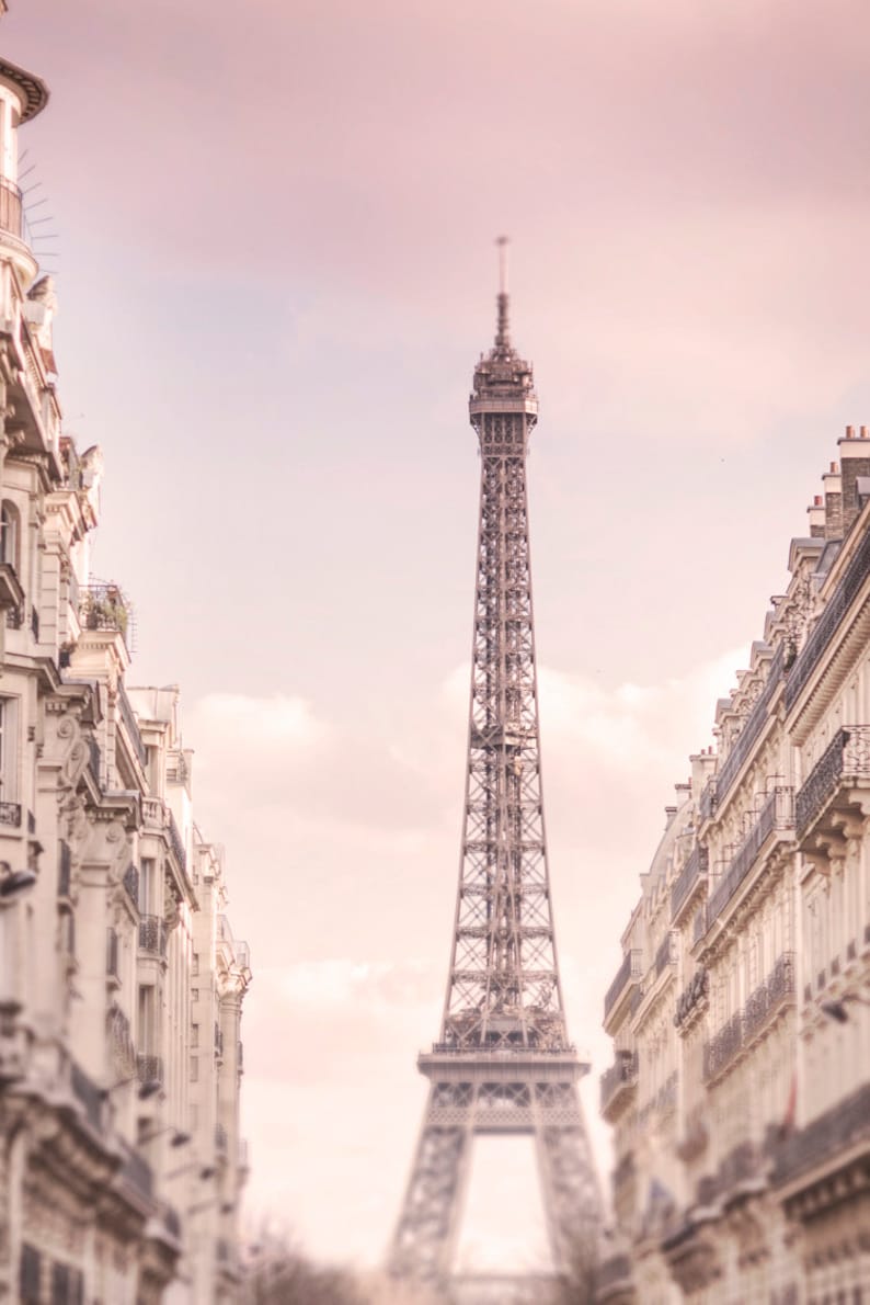Pode incluir: Fotografia da Torre Eiffel em Paris, enquadrada por edif&iacute;cios. A torre &eacute; castanho claro, o c&eacute;u &eacute; rosa p&aacute;lido. Os edif&iacute;cios s&atilde;o brancos com detalhes ornamentados.