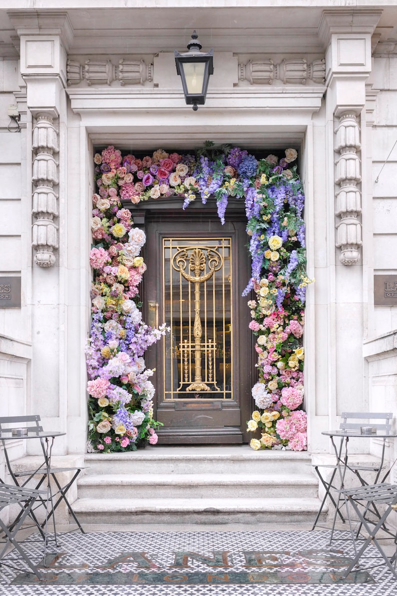 May include: A white building with a doorway decorated with a floral arrangement of pink, purple, and yellow flowers. The doorway has a dark wooden door with a gold metal grate and a black light fixture above it.
