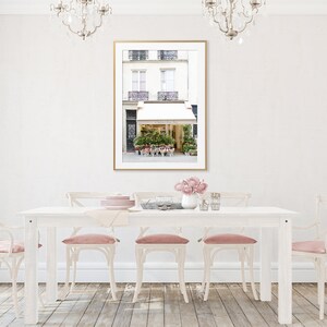May include: A dining room scene with a white table and chairs, pink cushions, and a framed picture of a Parisian flower shop. The room has hardwood floors, sheer pink curtains, and crystal chandeliers.