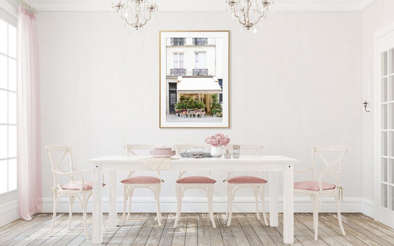 May include: A dining room scene with a white table and chairs, pink cushions, and a framed picture of a Parisian flower shop. The room has hardwood floors, sheer pink curtains, and crystal chandeliers.