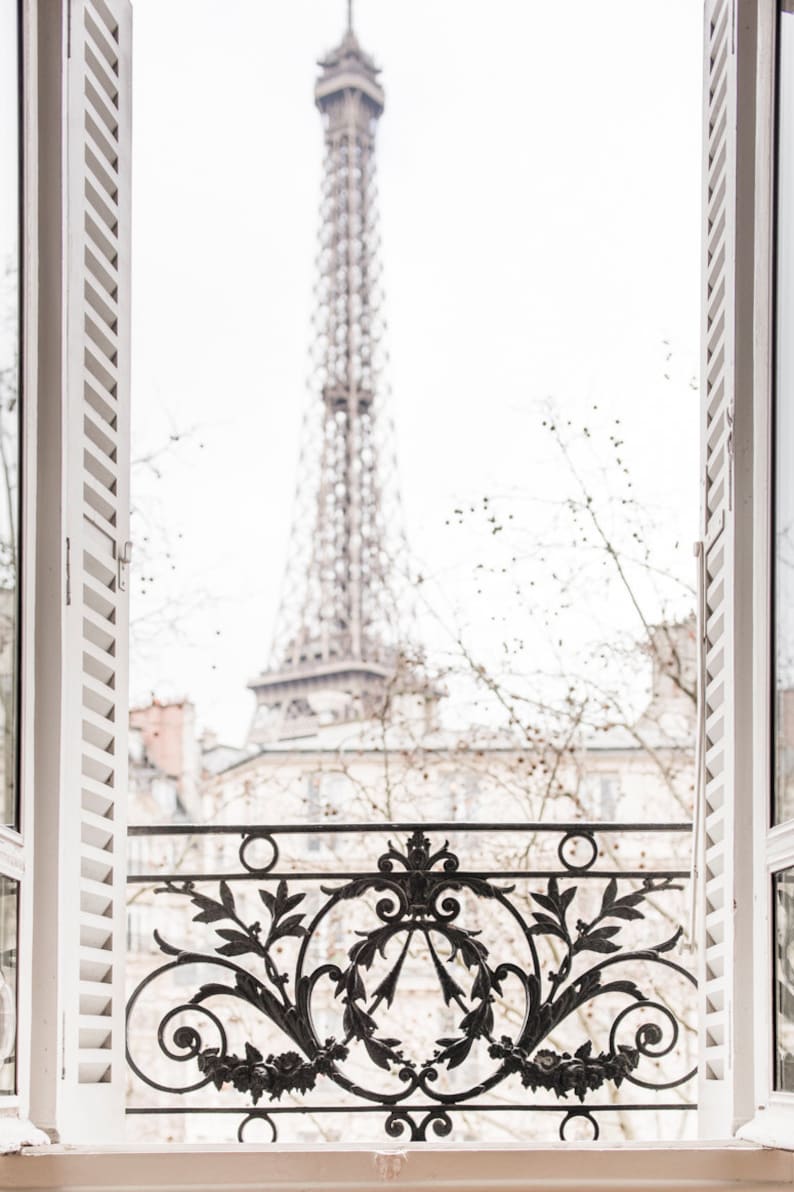 May include: A view of the Eiffel Tower in Paris, France, seen through a window with white shutters and a black wrought iron balcony railing.