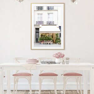 May include: A framed print of a Parisian flower shop with a gold frame hangs above a white dining table. The shop has a white awning and is filled with colorful flowers. Pink velvet chairs surround the table, and a vase of pink flowers sits on the table.