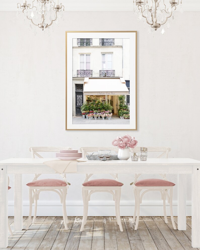 May include: A framed print of a Parisian flower shop with a gold frame hangs above a white dining table. The shop has a white awning and is filled with colorful flowers. Pink velvet chairs surround the table, and a vase of pink flowers sits on the table.
