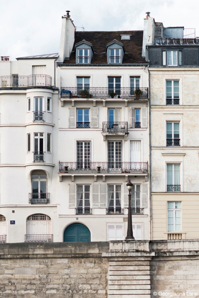 May include: A white building with a teal blue door and multiple windows. The building has a balcony with a wrought iron railing. The building is located in a city and has a stone wall in front of it.