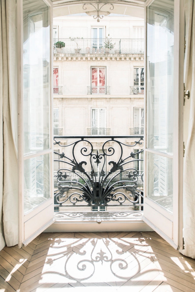 May include: A view from a window with white French doors, looking out at a building with a wrought iron balcony. The balcony is decorated with intricate scrollwork and floral designs. The building is painted in a light cream color and has several windows. The sun is shining brightly, casting a shadow of the balcony on the floor.