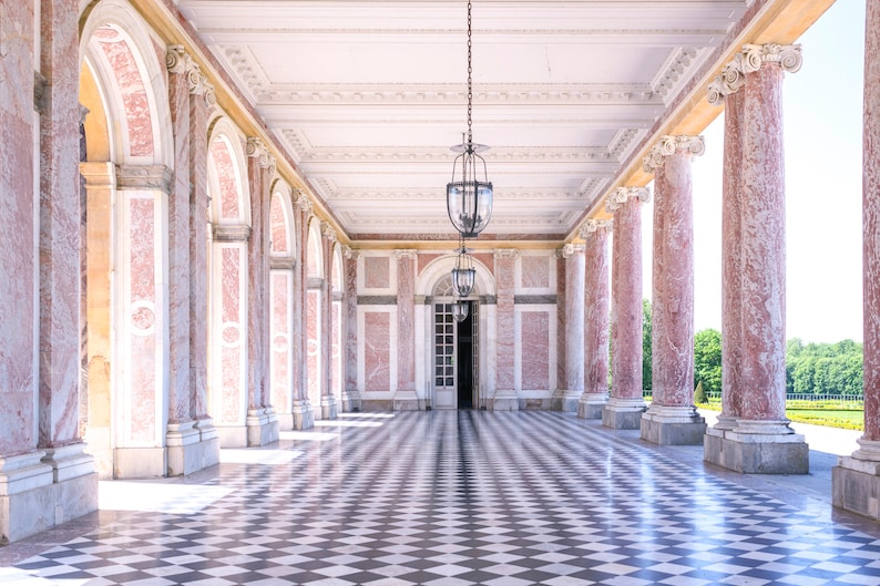 May include: An interior shot of a colonnade with pink marble columns and a checkered floor. The ceiling is white with ornate molding, and two hanging lanterns illuminate the space. A doorway is visible at the end.