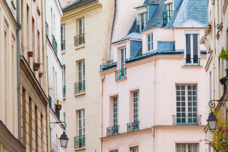 May include: A narrow street in Paris, France, with several buildings lining the sides. The buildings are all painted in shades of pink, white, and beige. The windows are all different sizes and shapes, and some have balconies. The street is empty, and the only light comes from the sun shining through the windows.