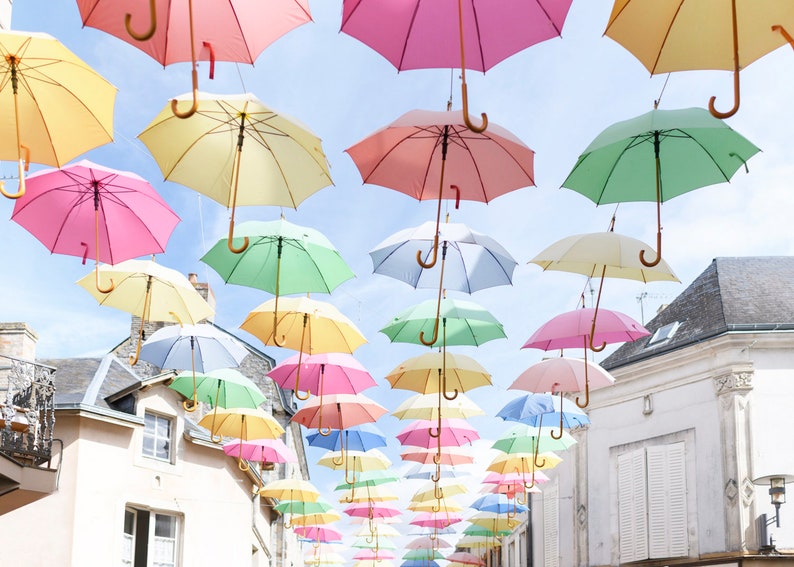 May include: A street lined with colorful umbrellas hanging overhead, creating a whimsical and cheerful atmosphere. The umbrellas are in various shades of pink, yellow, green, and blue, creating a vibrant display.