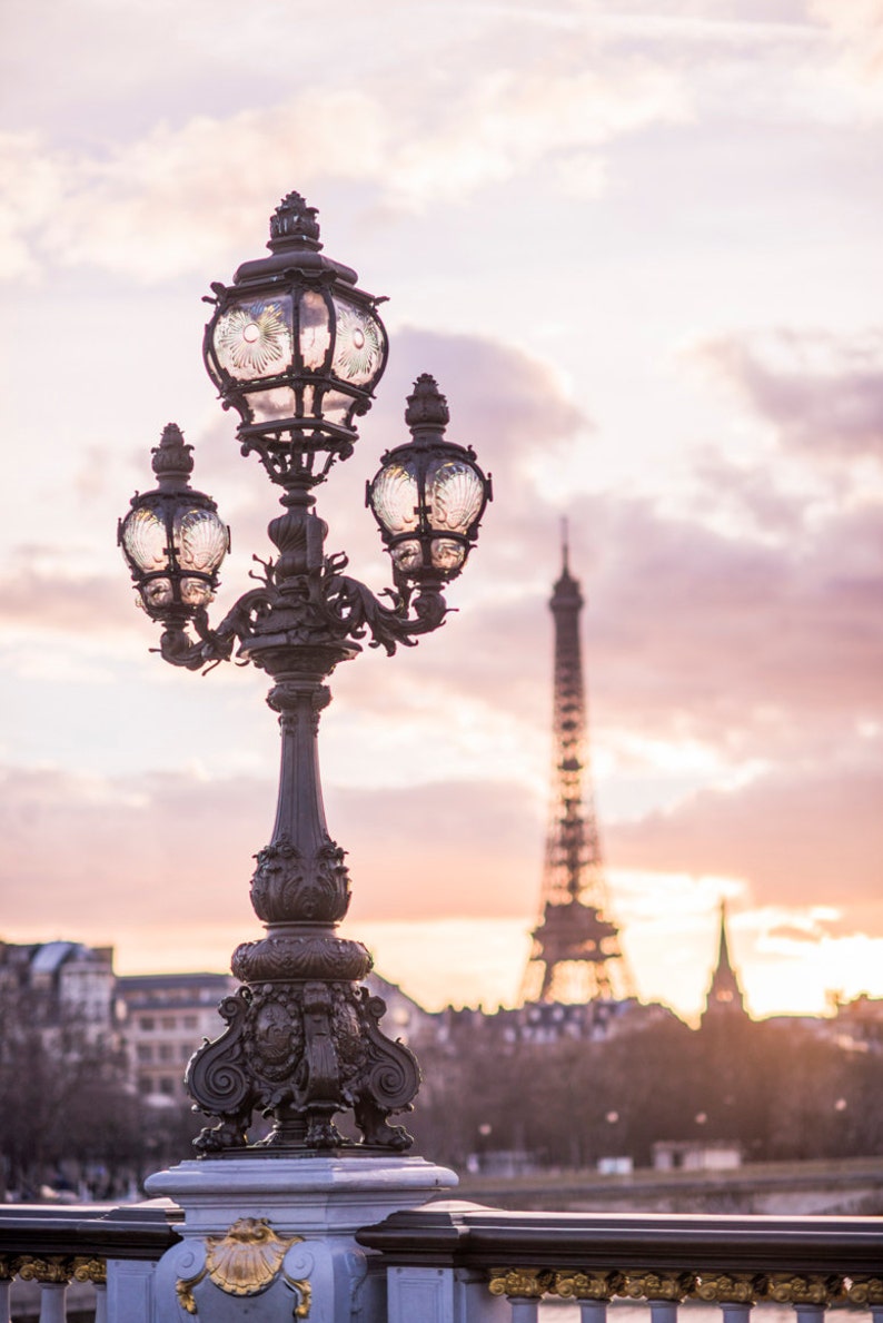 May include: A black wrought iron lamp post with three ornate glass globes stands on a bridge with the Eiffel Tower in the background. The sky is a soft pink and purple.
