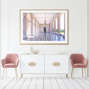 May include: A framed print of a long hallway with columns and a checkered floor. The print is above a white cabinet with gold hardware and two pink velvet chairs with gold legs. The room has white walls and a white floor.