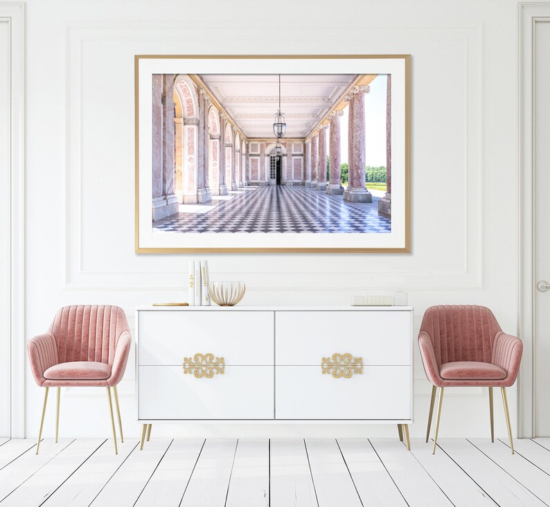 May include: A framed print of a long hallway with columns and a checkered floor. The print is above a white cabinet with gold hardware and two pink velvet chairs with gold legs. The room has white walls and a white floor.