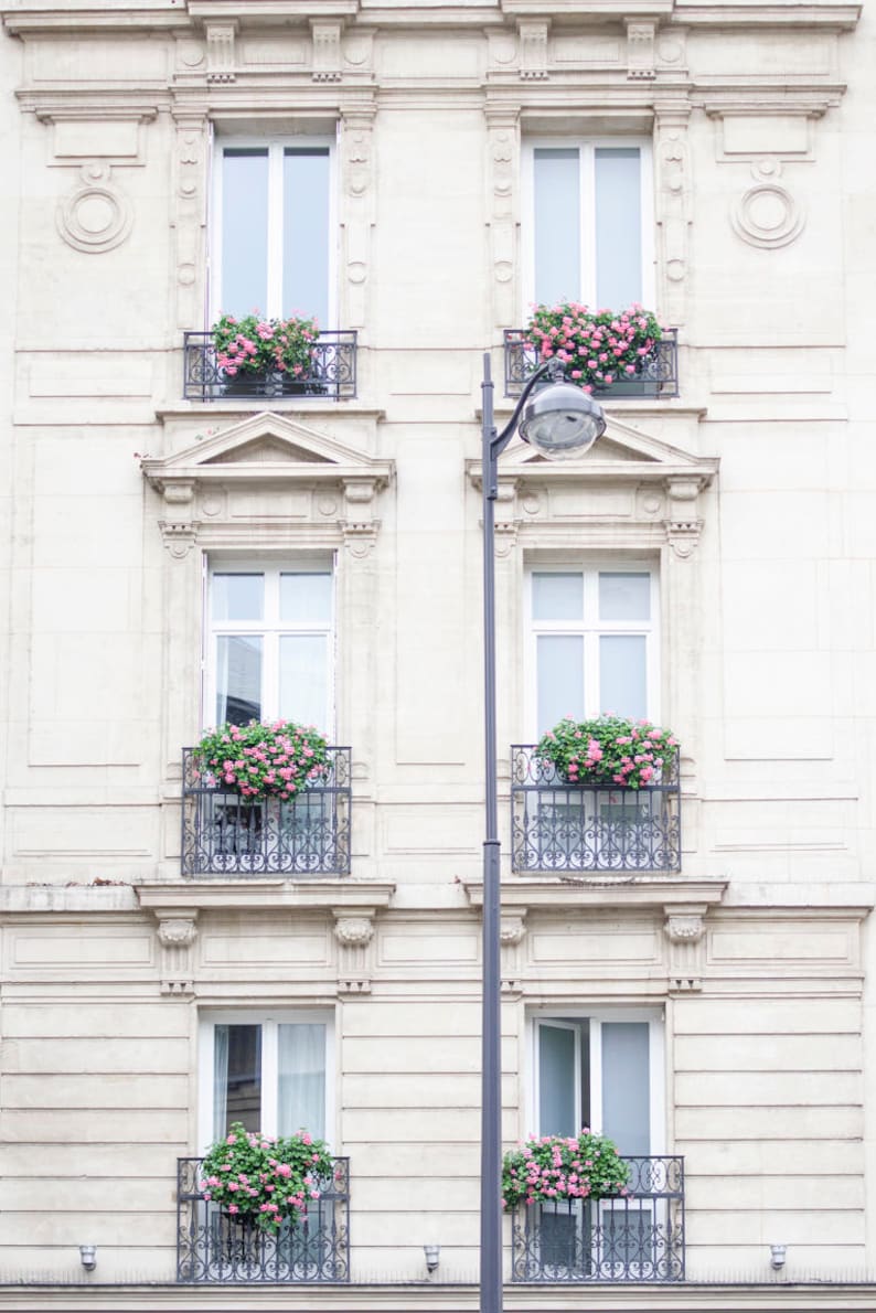 May include: A white building with four windows and balconies. Each balcony has a wrought iron railing and is decorated with pink flowers.