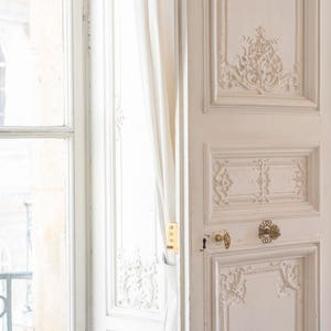 May include: A white door with intricate carvings and gold hardware. The door is open and a white curtain is partially visible behind it.