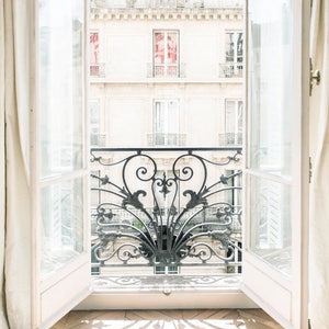 May include: A white door with glass panels opens to reveal a view of a balcony with an ornate wrought iron railing. The balcony is on a building with white walls and windows. Sunlight streams through the open door, casting a shadow of the railing on the wooden floor.