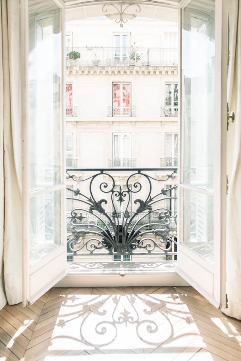 May include: A white door with glass panels opens to reveal a view of a balcony with an ornate wrought iron railing. The balcony is on a building with white walls and windows. Sunlight streams through the open door, casting a shadow of the railing on the wooden floor.