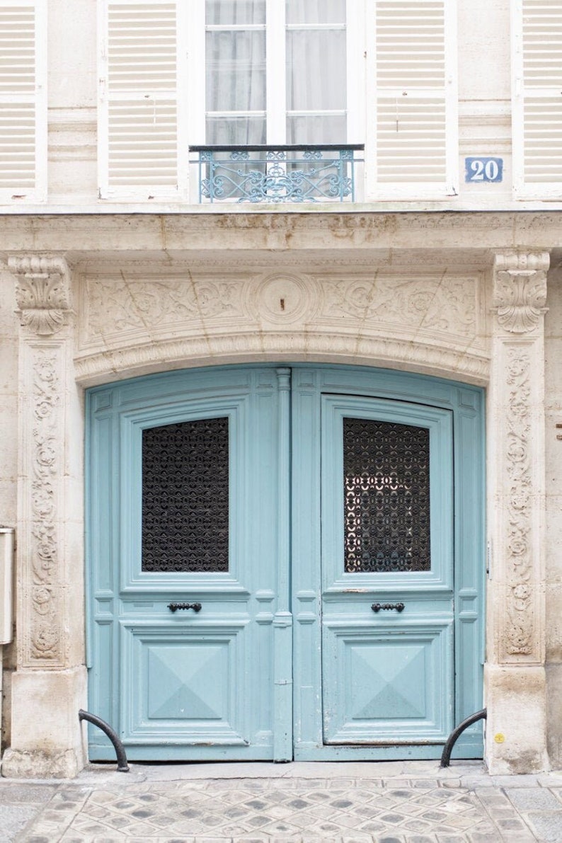 May include: A light blue double door with a decorative iron grate window above. The door has a simple design with a raised panel and a small window with a black grate. The door is set in a white stone archway with the number 20 on the wall above.