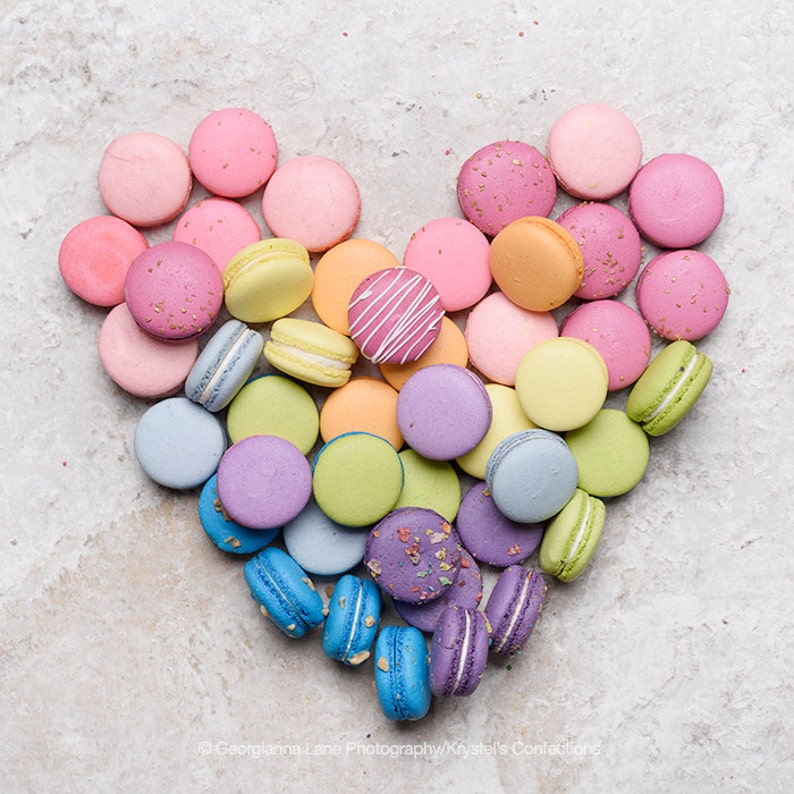 May include: A heart-shaped arrangement of colorful macarons, with shades of pink, orange, yellow, green, blue, and purple. The macarons are arranged in a gradient pattern, with the pink macarons at the top and the purple macarons at the bottom.