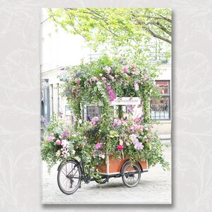 May include: A flower cart decorated with pink, purple, and white flowers. The cart is a bicycle with a wooden box on the back. The cart is parked on a cobblestone street. The sign on the cart reads 