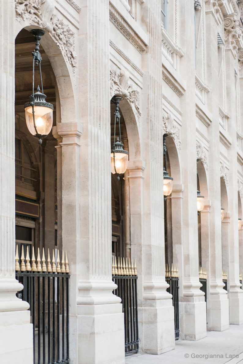 May include: A row of white stone arches with ornate details and black metal gates. Each archway has a hanging lantern with a warm yellow light.