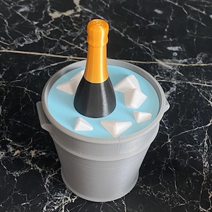 Può includere: Un secchiello per champagne grigio con interno azzurro, contenente una bottiglia di champagne nera e oro e cubetti di ghiaccio bianchi. Il secchiello ha due manici ed è su una superficie di marmo nero.