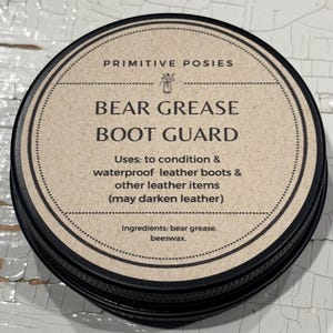 May include: A round, black tin of Primitive Posies Bear Grease Boot Guard. The label is tan with black text that reads "Bear Grease Boot Guard." It conditions and waterproofs leather boots and other leather items. Ingredients: bear grease, beeswax.