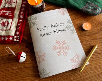 May include: A white planner with a snowflake pattern and the text "Family Activity Advent Planner". The planner is on a wooden surface with a red and white quilt, a green sock, and two lit candles.