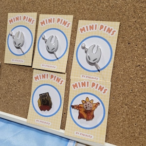 Peut inclure: Plusieurs mini-épingles sont présentées sur un tableau en liège. Les épingles présentent divers motifs, dont un personnage blanc stylisé, un livre avec le chiffre 3 et un personnage en robe orange. Chaque épingle est sur une carte avec "MINI PINS" et "BY HYRIAVEN".