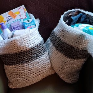 May include: Two white and grey crocheted bags filled with baby items. The bag on the left contains a book, bottles of lotion, and a nappy rash cream. The bag on the right contains a pack of nappies and a blue and white blanket.