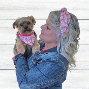 May include: A woman wearing a pink and white floral headband holds a small Yorkshire Terrier wearing a matching pink and white floral bandana.