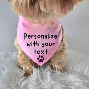 May include: A light pink triangular pet bandana featuring the black printed text "Personalize with your text" centered above a black paw print graphic. The fabric is smooth and shaped to drape around the neck of a small, furry animal.